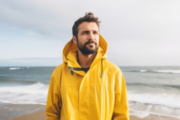 Portrait of a tender man in his 30s wearing a vibrant raincoat while standing against serene seaside background