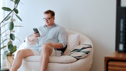 Middle age man sit on sofa with coffee cup in room using phone search data for occupation, raising...