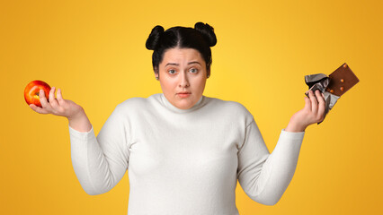 Puzzled Overweight Woman Holding Chocolate Bar And Apple In Hands, Can't Decide What To Choose. Yellow Studio Background, Panorama