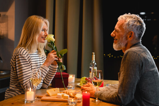 Happy middle-aged woman enjoying the rose fragrance during romantic dinner in the kitchen with her husband. Spouses wife and husband celebrating anniversary, special event, birthday, Valentine`s day
