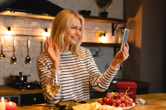 Cheerful mature caucasian woman having video call in the kitchen having romantic dinner. Vlogging and blogging for social media. Celebration and holiday
