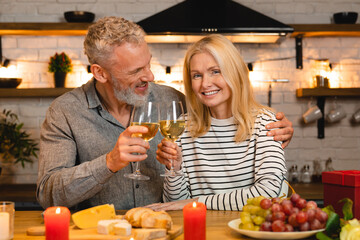Cheerful mature caucasian couple having romantic date in the kitchen while dining and drinking wine alcoholic beverage, celebrating anniversary, quality time together