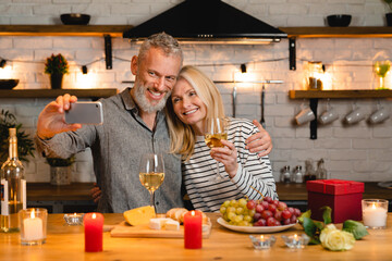Happy caucasian aged couple taking selfie using mobile phone on a romantic date in the kitchen. Celebration of special event, anniversary, engagement, proposal online on vlog blog
