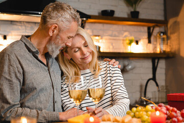 Passionate mature caucasian couple during romantic date in the kitchen. Loving husband spouse kissing cuddling embracing hugging his wife girlfriend while dining on special event, anniversary