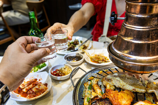 Two person toasting soju drinks during dinner in korean barbecue restaurant while enjoying tasty grilled meats.