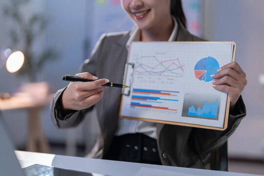 Businesswoman is pointing at charts on a clipboard with a pen while smiling. She is delivering a presentation in an office setting