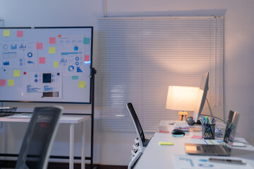 Empty modern office at night with a lit laptop on desk and whiteboard with business data