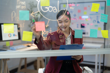 Young manager is using colorful sticky notes on a glass wall to plan her next project, writing down...