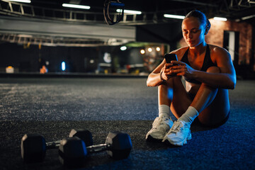 Young athlete woman sitting on gym floor browsing smartphone after workout session