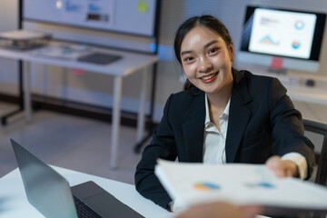 Smiling asian businesswoman hands paperwork to a colleague in a modern office, showcasing teamwork and communication