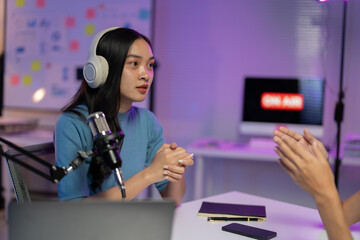 Asian woman hosts a podcast, interviewing a guest in her cozy home studio. She records with a microphone, showcasing modern content creation