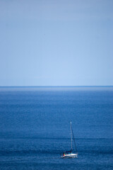 Fototapeta premium A small sailboat with a Portuguese flag floats on the calm waters of the Atlantic Ocean, taken off the coast of the Azores islands. The vast, clear blue sea stretches into the horizon.