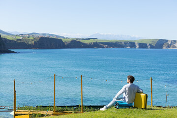 Fototapeta premium Single young adult man resting leaning on his yellow travel suitcase with the background of the sky and the ocean or sea, rest vacation, traveling the world, traveling alone 
