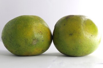 The photo shows two green oranges on a white background. The oranges are round and have a smooth skin. They are slightly different in size.