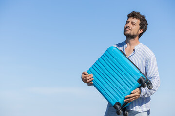 Happy and euphoric single young adult man with his blue travel suitcase with the background of blue...