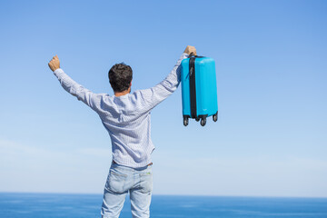 Happy and euphoric single young adult man with his blue travel suitcase with the background of blue...