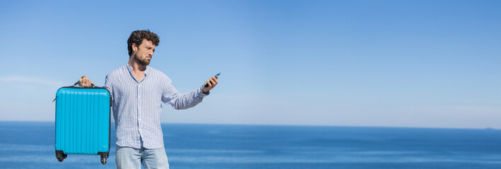 Happy single young adult man booking vacation hotel with his smartphone, blue travel suitcase with...