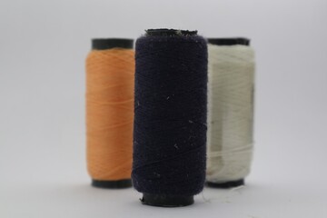 A close-up of three spools of thread against a white background. The spools are arranged in a row and are of different colors: orange, navy blue, and white.