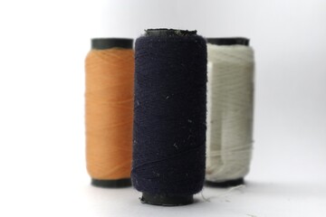 A close-up of three spools of thread against a white background. The spools are arranged in a row and are of different colors: orange, navy blue, and white.