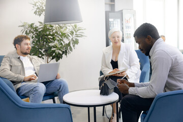 Diverse team engages in collaborative meeting in modern office space. two individuals are seated with laptops, while another takes notes, fostering productive work environment.