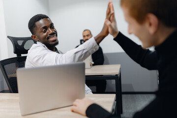 African american and caucasian businessmen in office setting engage in high-five, symbolizing team success and collaboration. colleague can be seen in background, enhancing atmosphere of teamwork.