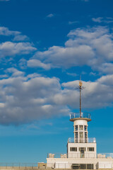 Obraz premium Upper part of a modern river station building with a glass observation deck and metal spire against a bright blue sky with scattered clouds. The structure is illuminated by sunlight. 