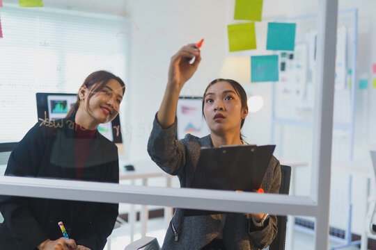 Two young businesswomen brainstorming and writing on a glass panel in their office - Powered by Adobe