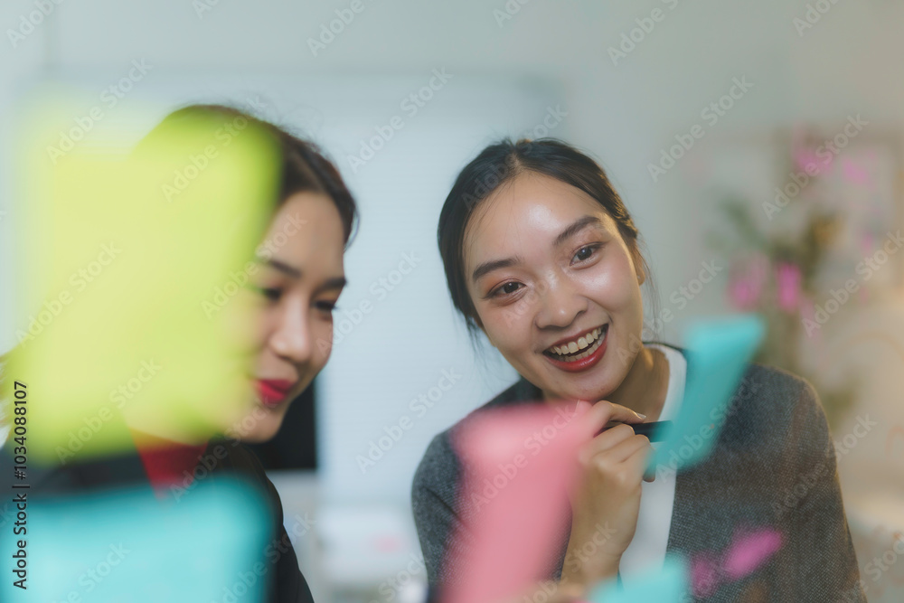 Wall mural two businesswomen are smiling while brainstorming and planning using adhesive notes on a glass wall 