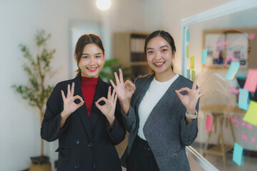 Two young businesswomen are smiling and showing ok sign with both hands while standing in the office