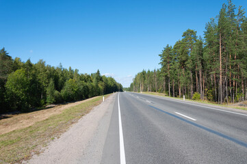 Fototapeta premium Asphalt highway through the forest in Karelia, blue sky, sunny summer day