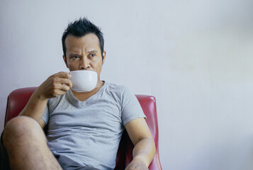 Asian man enjoying a cup of coffee mug sitting relaxed on sofa at home.