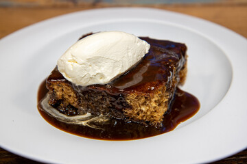 A white plate of delicious sticky toffee pudding chocolate cake with cream and chocolate sauce on a wooden kitchen table work top