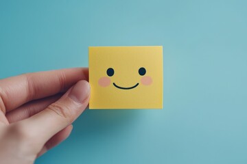Obraz premium A hand holding a cheerful yellow sticky note with a smiling face against a light blue background indoors during daylight