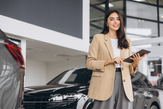 Smiling car seller, businesswoman in beige suit standing in car salon and using tablet for choosing right car. In background are many modern cars.