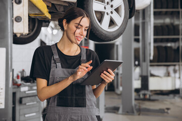 Young pretty smiling female mechanic, in grey uniform, holding tablet in her hands, poses standing under car on lift.