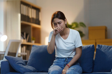 Young woman is sitting on her sofa at night, feeling unwell and coughing into her hand