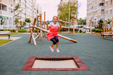 A girl in a pink tunic is jumping on a trampoline in the yard at a sports playground in the city