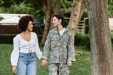 A female soldier in camouflage is joyfully welcomed home by her loving African American wife in a serene outdoor setting.