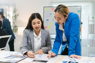 Two businesswomen are having a working meeting, standing and analyzing charts and business data on a desk