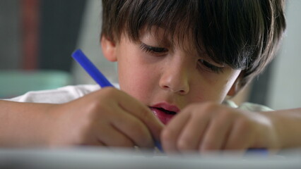 Close-up of young boy deeply focused on coloring with a blue marker, showcasing concentration and...