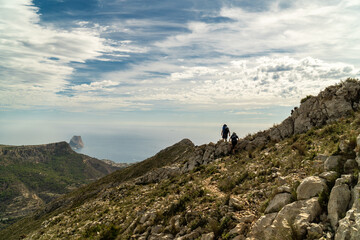 Silhouette of two hikers walking on a hill, against a cloudy sky, backlit.