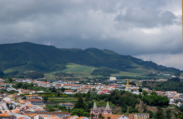 Fototapeta premium A scenic view of Angra do Heroísmo, a historic town on Terceira Island in the Azores, Portugal. The image shows the town's red-tiled roofs, lush green hills, and dramatic cloudy skies.