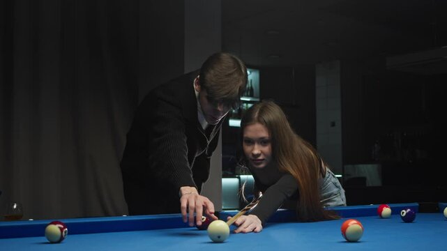 a young man teaches his girlfriend to play billiards. He explains the rules of the American pool game and guides her cue