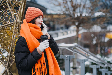 Millennial woman in cozy orange winter gear using smartphone. Christmas Urban tech-savvy style, winter warmth, city scene, hot coffee in hand.