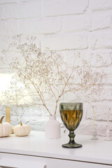  Home autumn composition.Charming green glassware with wine artfully combined with delicate decor of white pumpkins, lamp, dried branches on white bricks background.