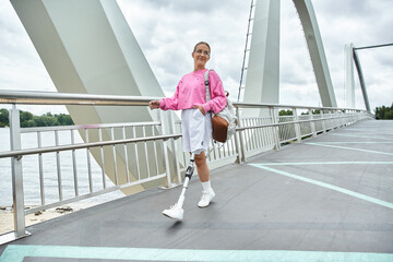 A young woman with a prosthetic leg walking on a bridge by the water.