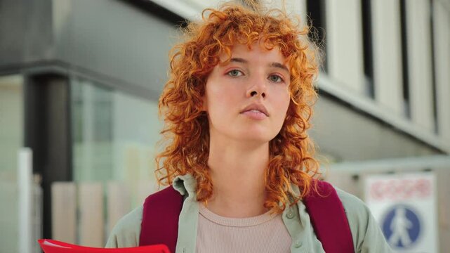 Young female student with red curly hair looking at the camera holding a bright red folder while standing outdoors on a modern campus. Thoughtful expression highlights focus and academic determination