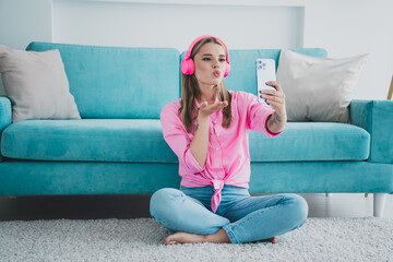 Young woman relaxing at home, blowing a kiss to her phone in vibrant headphones while enjoying...