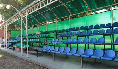 blue and green plastic chairs under a green canopy, a fragment of an open-air spectator stand without visitors, places for watching horse races at a provincial hippodrome