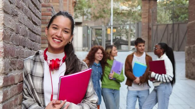 Young Hispanic female student smiling at the camera while holding a red notebook, standing against a brick wall on campus. A group of diverse students interacts in the blurred background outdoors.